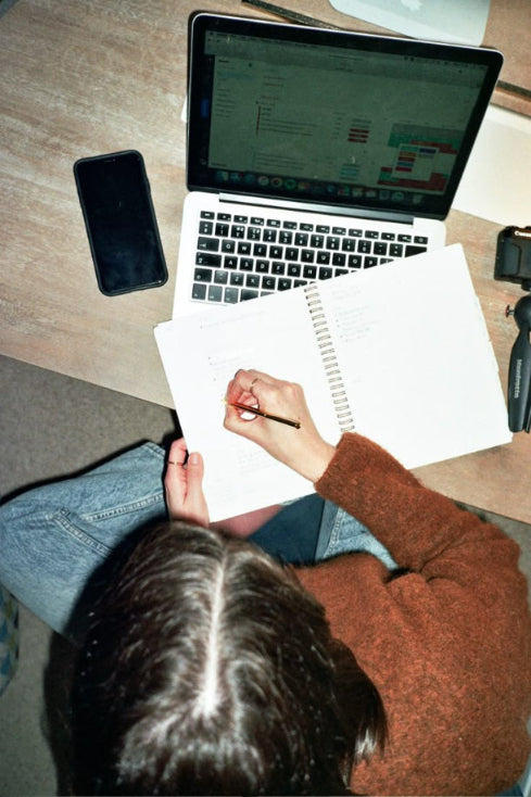 Person sitting at a desk with a laptop, phone, and notebook, wearing jeans and a brown sweater.