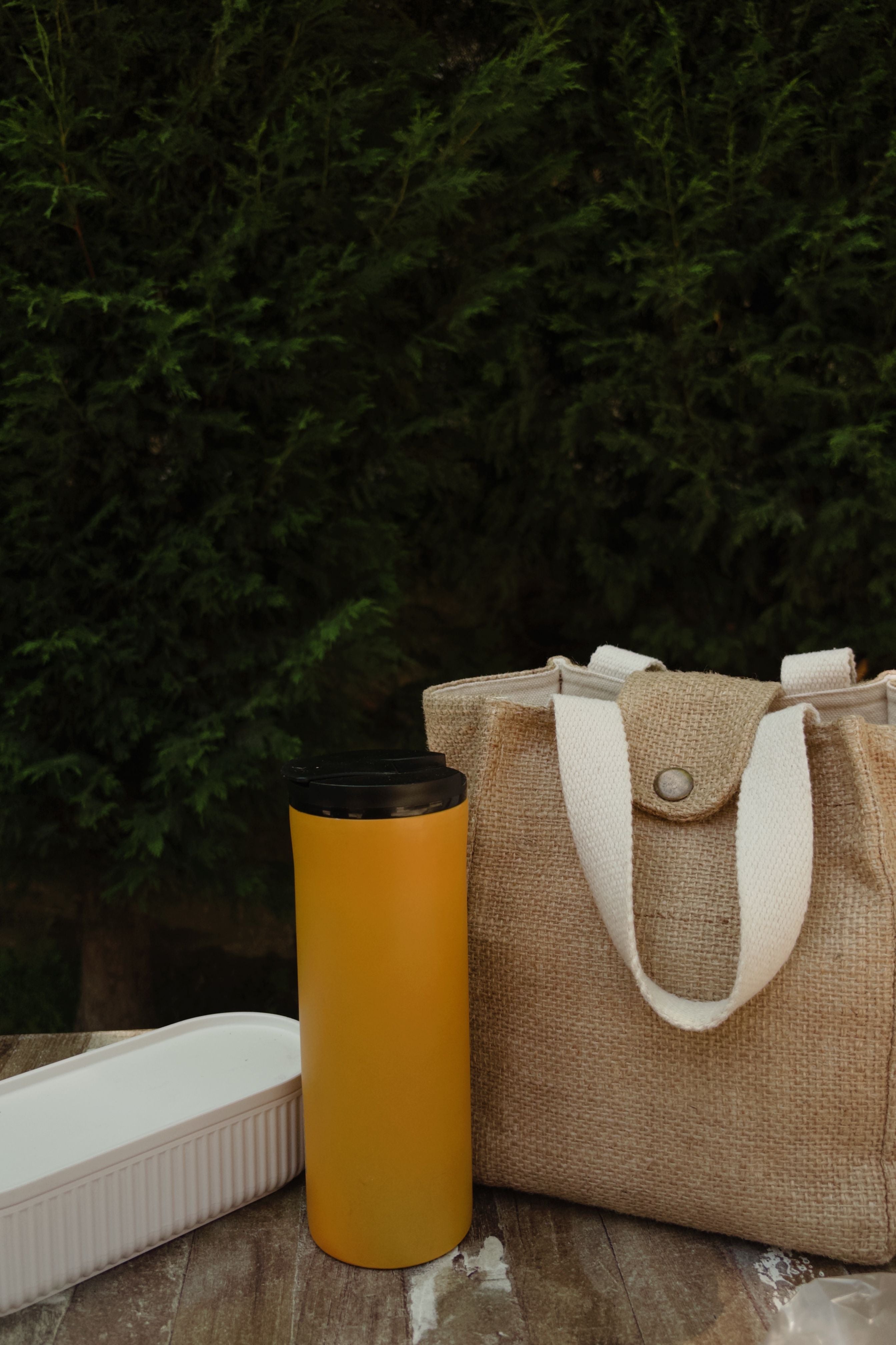 A yellow insulated travel tumbler with a black lid stands next to a natural burlap tote bag with white canvas handles on a wooden surface, with a white container and a dark green hedge background.